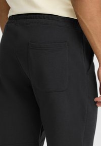 Un pantalon de survêtement noir avec une texture lisse, une taille élastique et une poche passepoilée à l'arrière, mettant en avant fonctionnalité et confort.