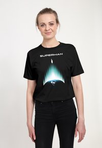 Žena v černém tričku Superman s potiskem Supermana létajícího vzhůru nad zemským obzorem na světlém pozadí.