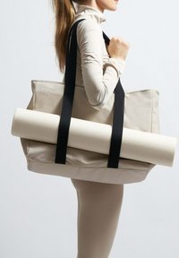 Béžová plátěná taška s černými popruhy, nese srolovanou bílou texturovanou jogamatku. Jednoduchý design s prostorným interiérem.