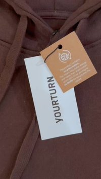 Hoodie de color marrón oscuro con capucha ajustable, una etiqueta con "TU TURNO" y una etiqueta de cuidado adjunta que detalla el contenido de algodón reciclado.