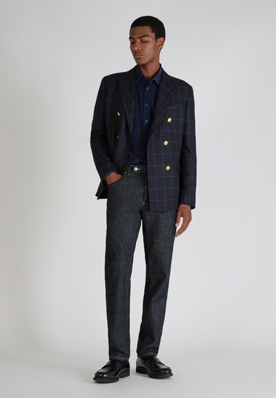 Dunkelblaues Karomuster-Doppelreiher-Blazer mit goldenen Knöpfen, kombiniert mit einem dunkelblauen Hemd und dunklen strukturierten Jeans, ergänzt durch schwarze Schuhe.