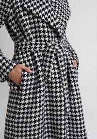 Płaszcz w czarno-białą kratę houndstooth z paskiem, z szerokim kołnierzem i bocznymi kieszeniami, wykonany z teksturowanej tkaniny.
