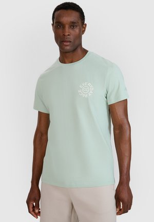 Man stands facing forward wearing a light green short-sleeve t-shirt with white "Everyday Pace" text on left chest and light pants.
