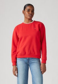 Red sweatshirt with a crew neck and white trim, featuring a small logo on the chest. Paired with light blue denim jeans.