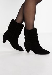 Mujer que lleva botas de ante negras hasta la mitad de la pantorrilla con un diseño arrugado y tacones bajos, combinadas con medias negras transparentes sobre un suelo blanco.