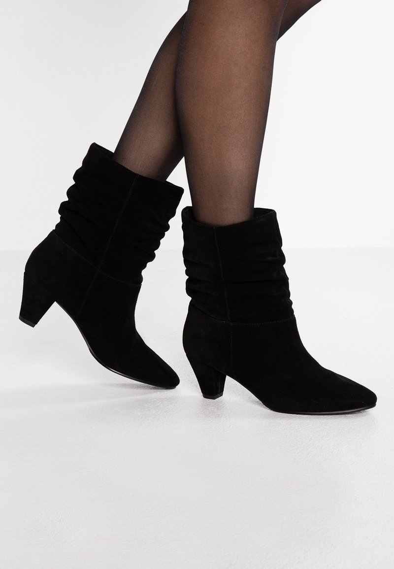 Mujer que lleva botas de ante negras hasta la mitad de la pantorrilla con un diseño arrugado y tacones bajos, combinadas con medias negras transparentes sobre un suelo blanco.