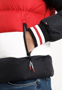 En person som bär en puffy-jacka i röd, vit och svart med en hand i den zipprade sidofickan och randiga ärmdetaljer.