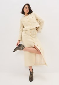 Ensemble deux pièces texturé couleur crème avec des accents de boutons dorés ; jupe à fente haute assortie à des bottines noir et doré à motif serpent.