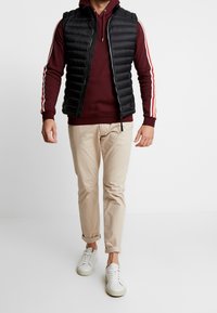 Black puffer vest over a burgundy hoodie with white and red stripes, paired with beige pants and white sneakers. Smooth materials, fitted design.