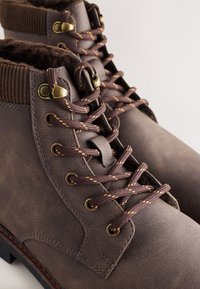 Smeđe gležnjače od teksturirane kože, s zlatnim metalnim obrubima za vezice, smeđim vezicama sa narančastim šavovima i rebrastim rubom za dodatnu toplinu.