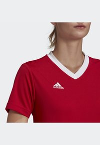 Punainen urheilupaita, jossa on valkoinen V-kaulus, lyhyet hihat ja valkoinen Adidas-logo vasemmassa rinta-alueessa. Sileä kankaan rakenne.