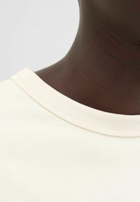 Camiseta de algodón de un beige claro con una textura suave y un cuello redondo acanalado, presentando un diseño simple sin patrones ni acentos.