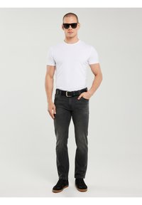 Mężczyzna w czarnych okularach przeciwsłonecznych, białej koszulce, czarnym pasku, szarych dżinsach i czarnych sportowych butach stoi na tle jasnoszarego tła.