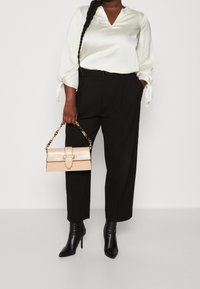 Cremefarbene Satinbluse mit Bindedetails, kombiniert mit schwarzen weit geschnittenen Hosen und schwarzen spitzen Stiefeln mit Absatz. Hält eine beigefarbene Handtasche mit goldenen Beschlägen.