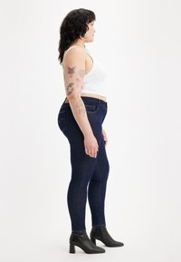 Mujer con cabello oscuro y múltiples tatuajes en los brazos, lleva una camiseta sin mangas blanca, jeans ajustados oscuros y botas de tacón negro hasta el tobillo, de pie de lado.