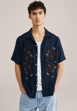 Marineblå kortærmet skjorte med blomsterbroderi i orange, blå og hvid. Lavet af lette materialer med en afslappet pasform og knaplukning foran.