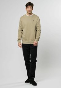 Felpa beige con colletto rotondo e maniche lunghe, polsini e orlo a coste. Jeans neri slim-fit e calzature nere completano il look.
