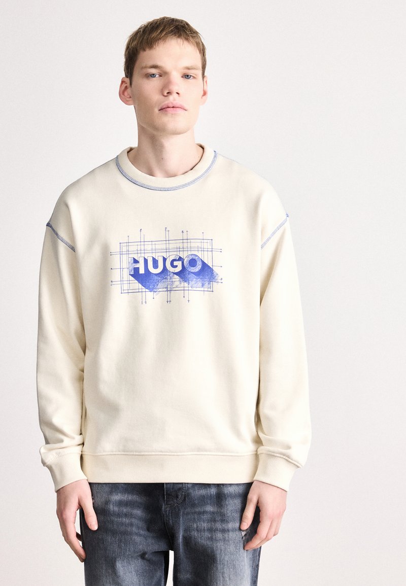 Krém színű pulóver bordázott mandzsettákkal, elöl kék grafikával, amely tartalmazza a "HUGO" szót és geometriai vonalakat.