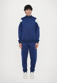 Tuta blu con giacca con zip e pantaloni abbinati. Presenta dettagli azzurri leggeri sulle spalle, polsini a coste e design sportivo. Sneakers bianche.