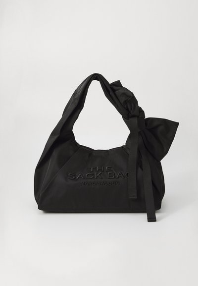 Sac en tissu noir avec une sangle nouée et un grand nœud décoratif. L'avant présente un texte en relief qui indique "THE SACK BAG MARC JACOBS."