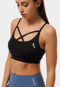 Melnā sporta krūšturis ar pārklājošām siksnām, izgatavots no elastīga auduma. Tam ir neliels balts logo kreisajā pusē.