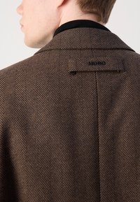 Barna herringbone mintás kabát fekete gallérral. Hátsó kihajtója logó részlettel rendelkezik. A texturált anyag mélységet ad a dizájnnak.