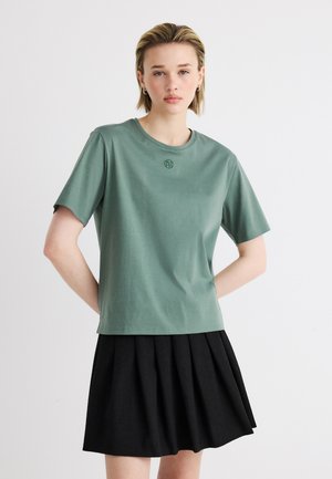 Young woman with short blonde hair wears a sage green short-sleeve top and a black pleated skirt against a plain background.