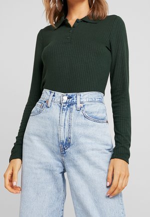 Kvinde iført en mørkegrøn ribbet langærmet poloshirt stoppet ned i højtaljede lyseblå acid-wash denimjeans.