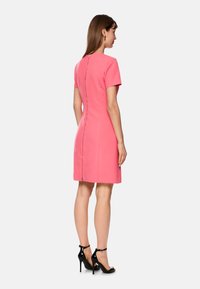 Kurzes pinkes Kleid mit kurzen Ärmeln, glatter Textur, geradem Schnitt und Reißverschluss hinten. Das Modell kombiniert es mit schwarzen High-Heels.