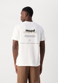 Balts kokvilnas T-krekls ar aizmugurējo grafiku, kas ir ar tekstu "mopd world.wide." Vienkāršs dizains, standarta piegriezums, īsas piedurknes, apgāzta apaļa kakla izgriezums.