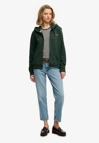 Sudadera verde con cremallera y dos bolsillos delanteros sobre una camiseta de rayas blancas y negras. Combinada con jeans azul claro y zapatos marrones sin cordones.