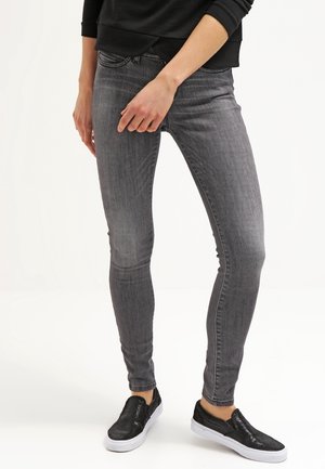 Jeans skinny grises con una textura suave y diseño de cinco bolsillos, combinados con zapatos negros tipo slip-on que tienen un upper texturizado y suela blanca.