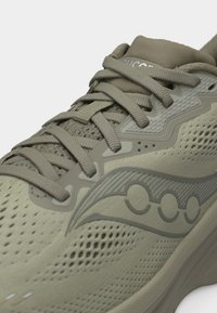 Κοντινό πλάνο ενός μπεζ αθλητικού παπουτσιού με υφαντό ύφασμα, υφές επικαλύψεων, μπεζ κορδόνια και λογότυπο σε σχήμα κύματος στην πλευρά.