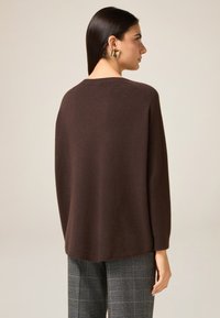 Brązowy, oversize'owy sweter o miękkiej fakturze, z prążkowanymi mankietami i zaokrąglonym dekoltem, w zestawie z kraciastymi spodniami. Prosty, casualowy design.