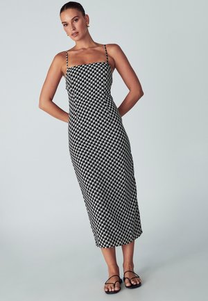 Čierno-biely vzorovaný midi šaty s tenkými ramienkami, priliehavým telom a rovnou siluetou, vystavené na svetlom pozadí.