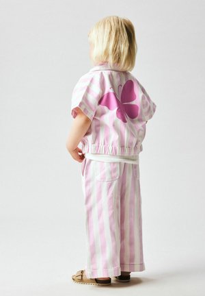 Laste blondide juustega, kes kannab roosa- ja valgetriibulist riietust, mille seljal on suur roosa liblikas, seisab seljaga vaataja poole.