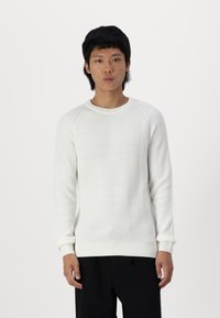 Maglione bianco a coste con scollo rotondo e maniche lunghe, caratterizzato da un knitting testurizzato. La vestibilità è leggermente ampia, abbinato a pantaloni neri.