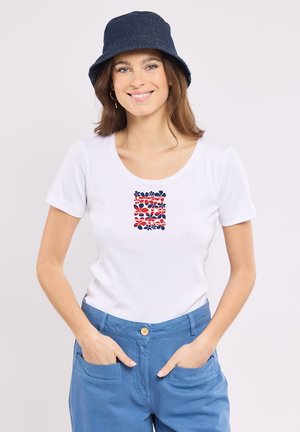 Femme souriante portant un t-shirt blanc avec un motif floral rouge et bleu marine, un pantalon bleu et un chapeau bob bleu marine, mains dans les poches.