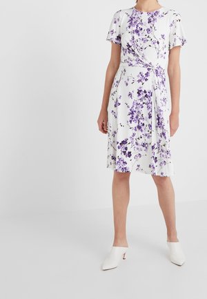 Woman wearing a white knee-length dress with purple floral print, short sleeves, side tie detail, and white heeled mules.