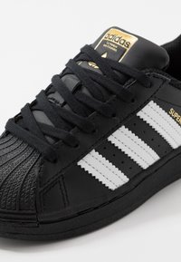Sneaker in pelle nera con punta testurizzata, caratterizzata da tre strisce bianche, lacci neri e un logo dorato Adidas sulla linguetta.