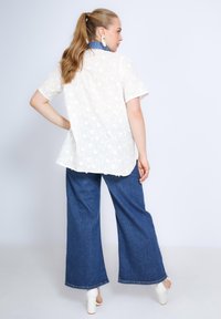 Witte bloemenkant korte mouwen top gecombineerd met blauwe wijdgerande denim broek en witte blokhak schoenen, die een relaxed fit en casual stijl tonen.