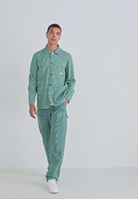 Light green denim jacket with a pocket, paired with matching cargo pants. Both pieces feature a subtle striped texture. White sneakers complete the look.