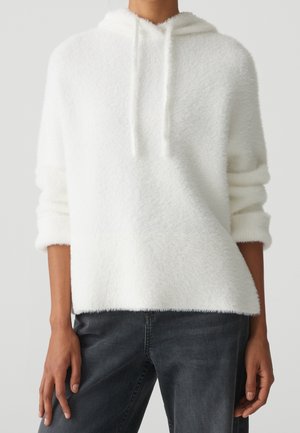 Personne portant un sweat à capuche blanc pelucheux avec des cordons et des manches retroussées, associé à un jean gris foncé, debout devant un fond uni.