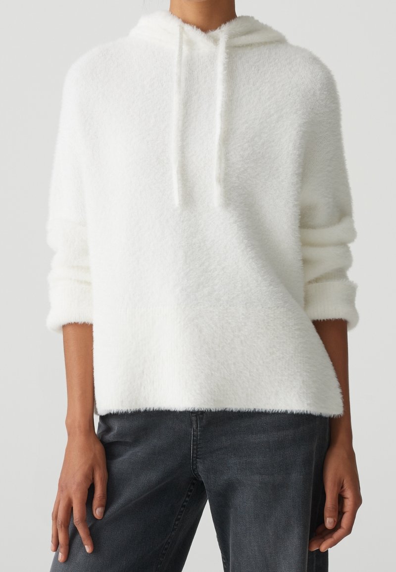 Personne portant un sweat à capuche blanc pelucheux avec des cordons et des manches retroussées, associé à un jean gris foncé, debout devant un fond uni.