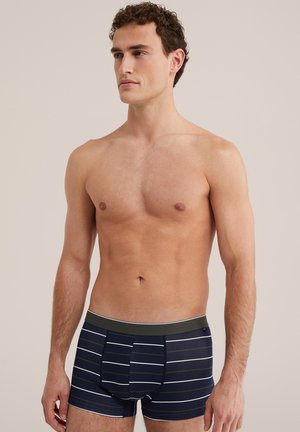 Mężczyzna bez koszulki z krótkimi kręconymi włosami, noszący granatowe bokserki z poziomymi białymi paskami na jednolitym jasnym tle.