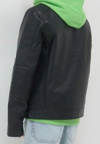 Czarna kurtka z ekoskóry z poziomymi żebrowanymi detalami na ramionach, noszona z zielonym dresowym swetrem z kapturem pod spodem.