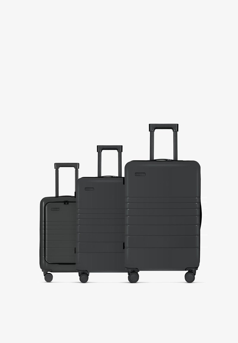 Three black hard-shell suitcases of varying sizes, featuring textured grooves, extendable handles, and multi-directional wheels.