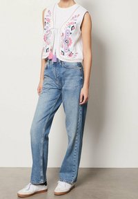 Biały, haftowany top bez rękawów z wiązaniem z przodu i różowymi frędzlami, zestawiony z luźnymi niebieskimi dżinsami i białymi trampkami.