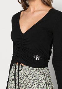 Black long-sleeve top with a V-neck and gathered front, featuring cinched ties at the hem and a small Calvin Klein logo.