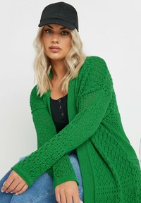 Grön stickad cardigan med öppen framsidan, texturerat diamantsmönster och långa ärmar. Kombinerad med en svart keps och svart topp. Jeansen i denim är synliga.